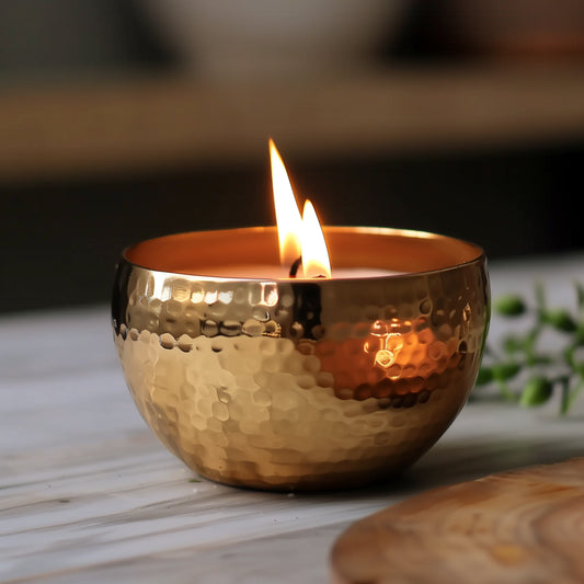 A lit brass vessel candle with a crackling wood wick, casting a warm glow on a marble surface in a cozy room setting.