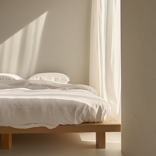 A minimalist bedroom with an organic cotton sheet set on a mattress on the floor, bathed in natural light from an open French window.